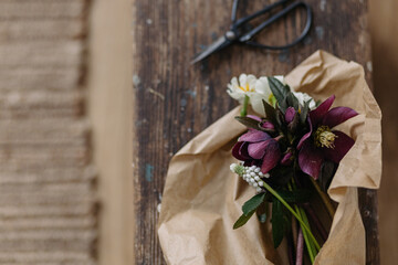 Beautiful spring bouquet in craft paper and scissors on rustic wooden background. First spring flowers helleborus, muscari, daffodils. Top view