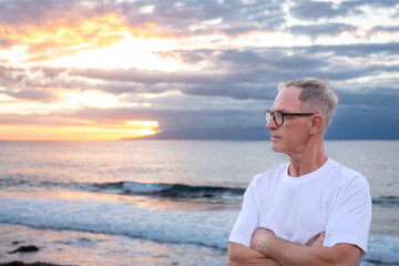 Fototapeta na wymiar Senior mature man at the sea beach at sunset light looking the horizon over sea admiring the orange sunray and cloudy sky, handsome man enjoys a relaxed retirement lifestyle