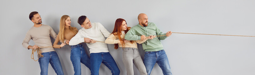 Group of happy adult friends playing tug of war and applying teamwork and unity strategy. Team of cheerful confident people standing together by grey wall pulling rope in one direction. Header, banner - obrazy, fototapety, plakaty