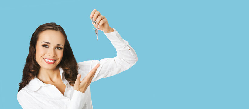 Cheerful smiling young businesswoman or real estate agent showing keys from new house. Portrait of excited brunette woman at studio, isolated aqua blue marine background with copy space. Wide image.