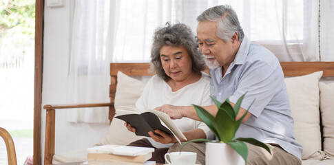 Happy Asian senior couple reading book together on couch in living room, Elderly woman laughing,...