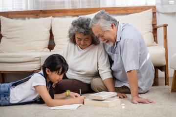 Asian senior couple watching granddaughter doing homework on the floor. Grandparents love and support little girl. Healthy living and lifestyle insurance - obrazy, fototapety, plakaty