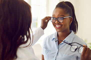 Optometrist visit. Joyful young dark-skinned woman is trying on new glasses she wants to buy in optical shop. Female optician gives African American female customer selection of eyeglasses. - obrazy, fototapety, plakaty