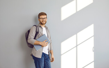 Portrait of smiling male adult student with backpack and folders over grey wall background. Bearded...