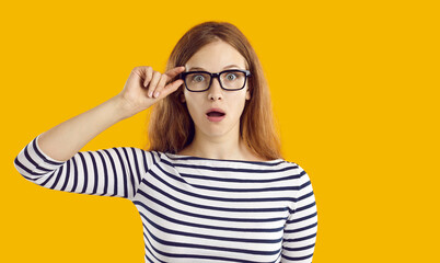 Young student girl very surprised by something. Beautiful nerdy young woman in striped top and glasses isolated on yellow background looking at camera with funny, shocked, astonished face expression - obrazy, fototapety, plakaty