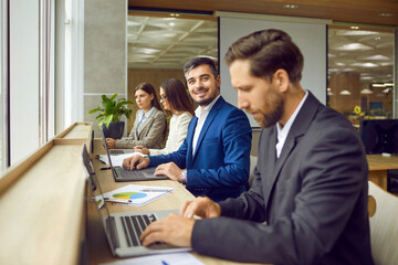 Positive businessman on workplace, clad in suit, sits at desk surrounded by the colleagues in the...