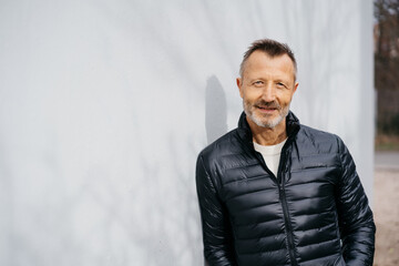 Confident Bestager Man in Stylish Black Jacket Leaning Against a Gray Concrete Wall with Copy Space