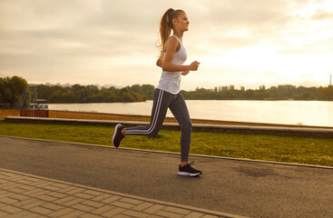 Cheerful slim girl athlete is exercising during morning run in fitness outfit. Smiling beautiful Caucasian young woman in sports tank top, leggings and sneakers jogging in park. Body care concept.