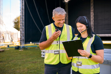 Male And Female Production Team With Headsets Setting Up Outdoor Stage For Music Festival Or Concert