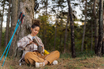 Sitting by the forest's border, the young woman with curly hair enjoys a peaceful coffee break while her trusty yellow backpack stands by.