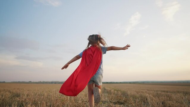 girl with the cloak. concept of a happy childhood and family for a child. a little girl in a superhero costume runs across a field, a red cape and a blue T-shirt, sunset lifestyle on the background
