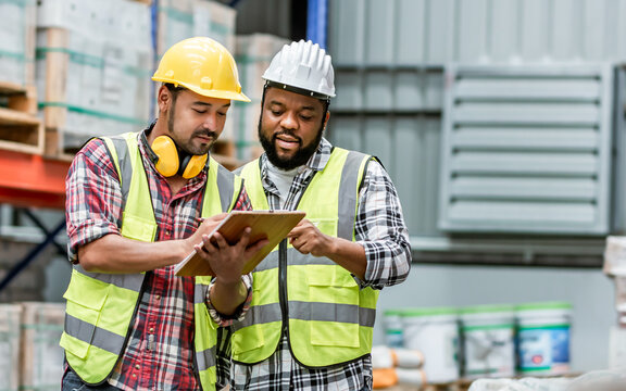 Two diverse professional male workers holding board, checking shipping stocks in storage, warehouse or factory for delivery, wearing safety hat. Diversity, Commercial Industry Business Concept.