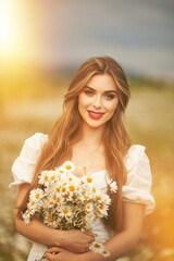 close portrait of a beautiful, laughing woman in a light dress holding a chamomile near her face during sunset, lit from the back