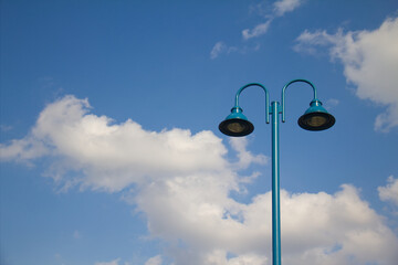 Teal blue metal street lamp stands against fluffy white clouds and bright blue sky.