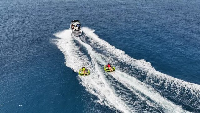 Aerial drone video of extreme inflatable towable tube cruising in high speed attached on powerboat with children on sofa ride, water sports in tropical exotic bay with deep blue crystal clear sea