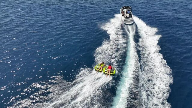 Aerial drone video of extreme inflatable towable tube cruising in high speed attached on powerboat with children on sofa ride, water sports in tropical exotic bay with deep blue crystal clear sea