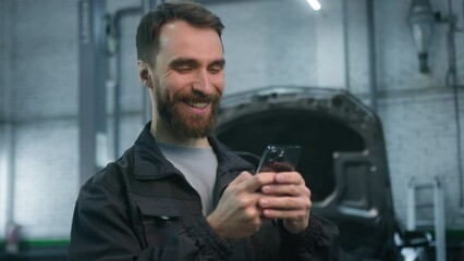Smiling cheerful man 30s Caucasian guy auto mechanic technical factory industry worker engineer in uniform using mobile phone looking at smartphone screen laughing work repair service at car garage