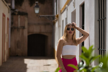 Obraz premium South American woman, young, beautiful and blonde with white top, pink pants and sunglasses, posing leaning on a white wall, in a sensual and provocative attitude. Concept trend, beauty, fashion.