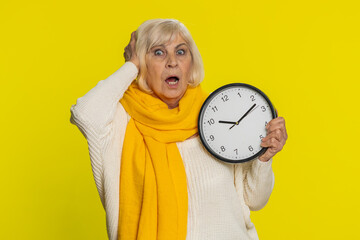 Senior old woman with anxiety checking time on clock, running late to work being in delay, deadline. Mature grandmother looking at hour, minutes, worrying to be punctual isolated on yellow background - obrazy, fototapety, plakaty