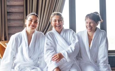 Three young smiling women in white bath robes on spa procedure. Attractive female friends laugh and relax during a skin care treatment.