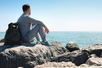 Caucasian guy in a blue sweater and hat holds his backpack and looking at the calm sea