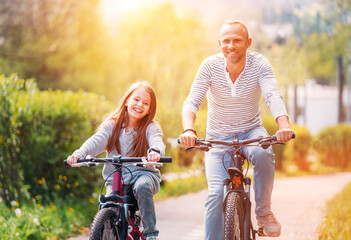 Portraits Smiling father with daughter during summer outdoor bicycle riding. They enjoy togetherness in the summer city park. Happy parenthood and childhood or active sport life concept image.