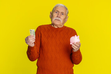 Poor senior old man insufficient amount of money, holding piggybank and one dollar banknote. Financial crisis. Bankruptcy. Poverty and destitution. Mature grandfather pensioner on yellow background