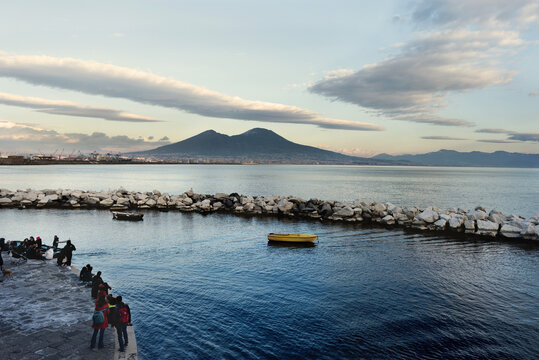 il Vesuvio visto dal lungomare di Mergellina, prospettiva con persone, barche e scogliera sullo sfondo, mare blu, e nuvole in movimento, vulcano dei campi flegrei, bellezza della natura, Napoli  