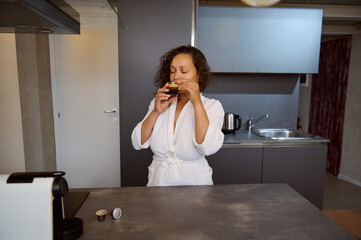 Fototapeta na wymiar Authentic portrait of a pretty woman in white bathrobe, enjoying her morning coffee, standing at kitchen counter