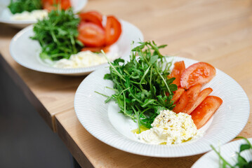 Fresh Arugula Salad with Tomato and Cheese. A healthy plate of green arugula salad paired with ripe tomato slices and a dollop of soft cheese, seasoned and ready to serve.