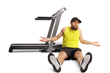 Confused bearded man in sportswear sitting on the floor in front of a treadmill