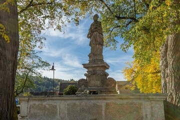 Obraz premium Svojsin, Czech Republic - October 13 2023: View of the stone water fountain with Johannes Nepomuk statue surrounded by yellow and green leaves. Sunny autumn day with blue sky.
