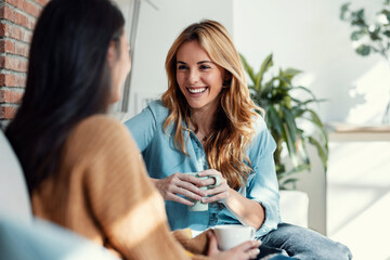 Two smiling young women talking while drinking coffee sitting on couch in the living room at home.