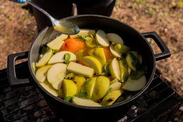 Glistening honey drizzles from a spoon into a pot of mulled wine with apples, oranges, and mint on a charcoal grill