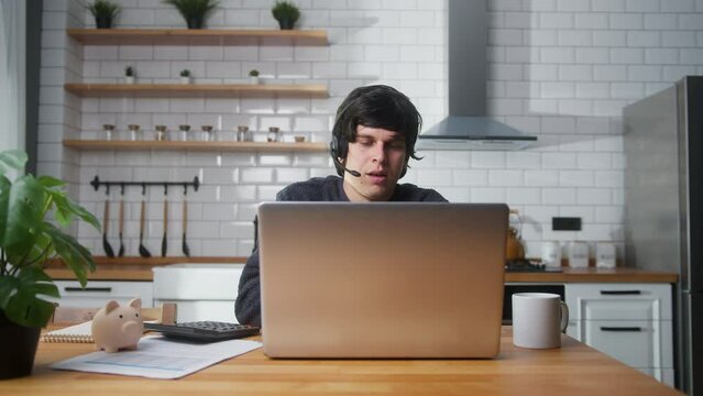 Focused young business man wear wireless headset sitting in the kitchen at home having video conference, typing on laptop, using calculator
