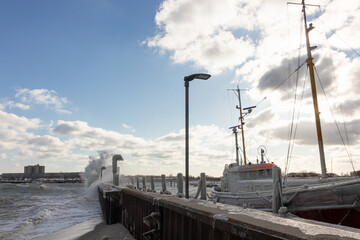 Große Welle klatscht gegen die Kaimauer am Hafen vom Küstenort Strande mit vereistem Fisch Kutter beim Wintersturm.