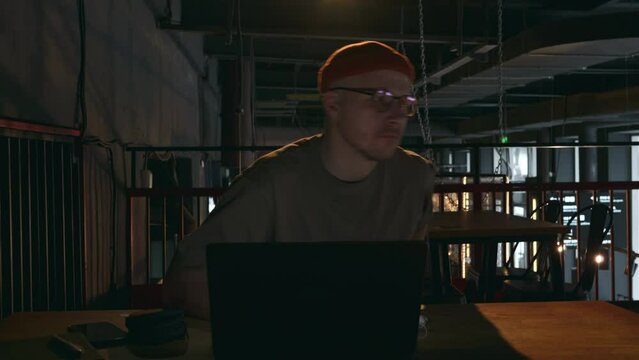 The cybercriminal is concentrating on his laptop in a dark room, then gets up and leaves his workplace. A young man is a millennial hipster hacker. High quality 4k footage