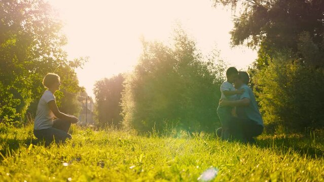 Squatting father warmly embraces son running with joy in field. Satisfied mother father and son create precious memories together in fresh air. Little boy hugs father running from mother on vocation