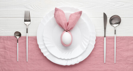 Easter holiday table setting with plate and cutlery, invitation concept. White wooden background. Bunny made of pink linen napkin and egg. Top view. Brunch or dinner.