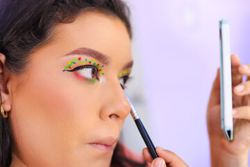 Close-up of a young Latina woman looking at herself in a small mirror while applying makeup, surrounded by a vibrant and colorful decoration