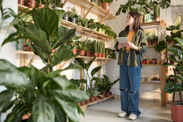 Fototapeta na wymiar Plant shop worker with tablet in her hands standing near shelves with plants and looking at them