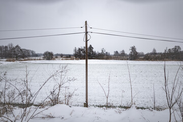 Ścieżka elektryczna idąca przez zaśnieżone pole porośnięte krzakami w popołudniowych godzinach pochmurnego szarego dnia w Zachodniej Polsce - obrazy, fototapety, plakaty
