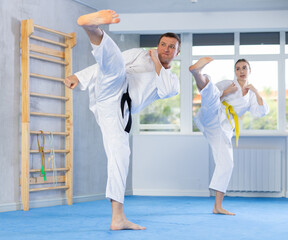 Dynamic young female attendee of karate classes and middle-aged trainer performing feet kicking technique in sports hall