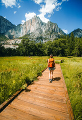 Fototapeta na wymiar Young woman wearing a backpack is walking on a wooden trail towards the Yosemite falls near Merced river in Yosemite valley, Yosemite National Park in United states of America