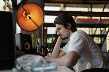 A focused man reviews content on his computer in a well-equipped home studio with a warm ambient light