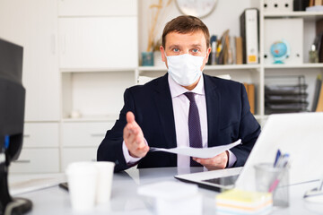 Confident manager in disposable surgical mask discussing financial report or explaining task while sitting in office. Concept of new life reality and precautions in COVID 19 pandemic