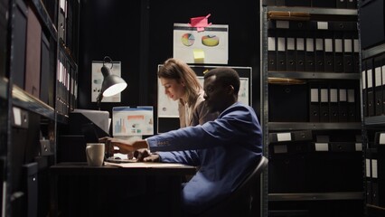 Diverse investigators studying the evidence in neatly organised office encircled by file shelves. Two law enforcement officials are carefully looking into criminal profiles and records on a laptop. - obrazy, fototapety, plakaty