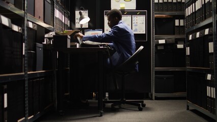 African American detective in archive room, reviewing evidence and files, with reliance on technology. Police officer conducts forensic investigation and background checks, ensuring justice shines. - obrazy, fototapety, plakaty