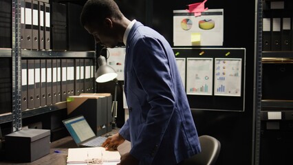 Devoted private investigator pulls files from shelf to conduct a comprehensive investigation. With laptop and research paperwork, african american agent analyzes evidence and gathers information. - obrazy, fototapety, plakaty