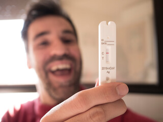 negative covid test is held in his hand by a man with black hair and a short beard, out of focus, happy with his mouth open, looking ahead at the test at home in front of the window. - 706005005